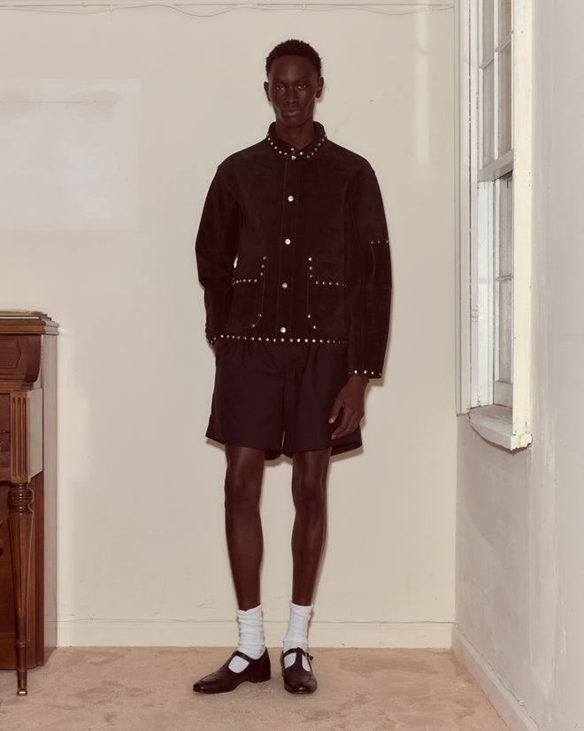 A person stands indoors wearing a Studded Welding Jacket - Black, paired with black shorts, white socks, and black shoes.
