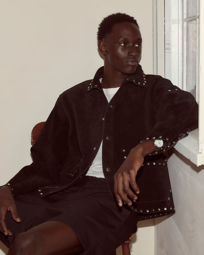 Wearing a black Studded Welding Jacket, a person sits by the window in shorts, gazing thoughtfully to the side.