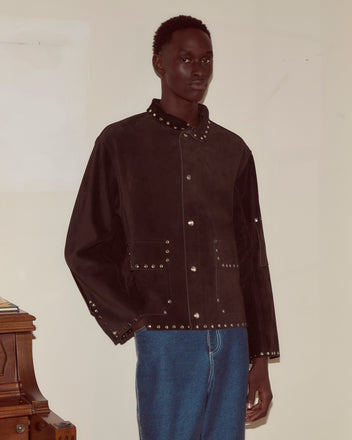 A person in a brown Studded Welding Jacket and blue jeans stands indoors against a plain wall.