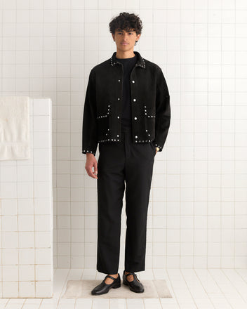 A person wearing the Studded Welding Jacket - Black, along with black pants and black shoes, stands in a white tiled room.
