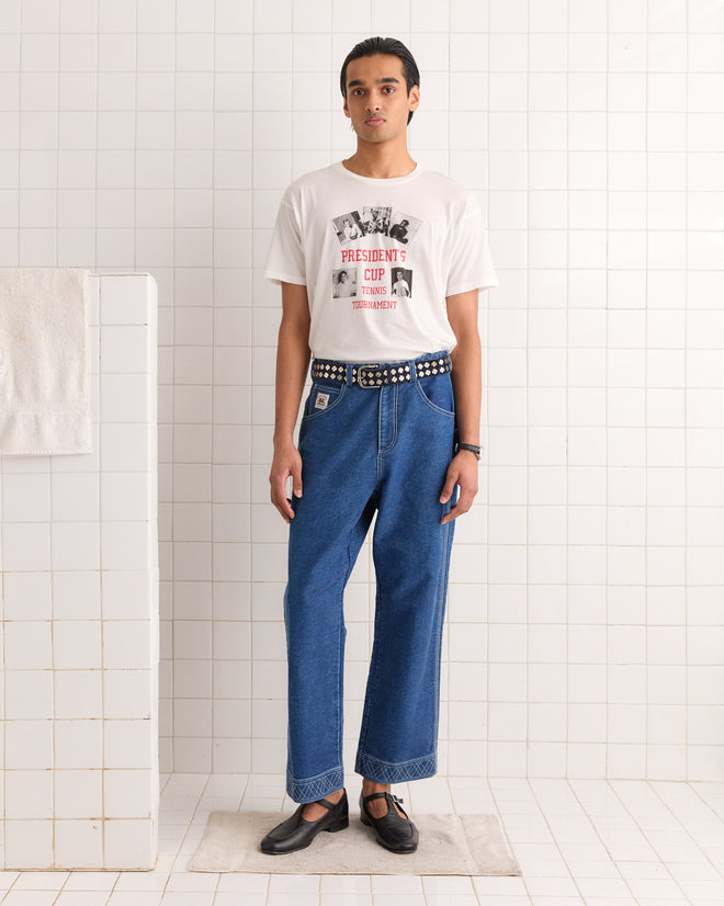 A person in a printed white t-shirt, blue jeans, and black shoes stands in a tiled room, with their look subtly enhanced by the vintage-inspired Clover Charm Belt in Dark Navy.