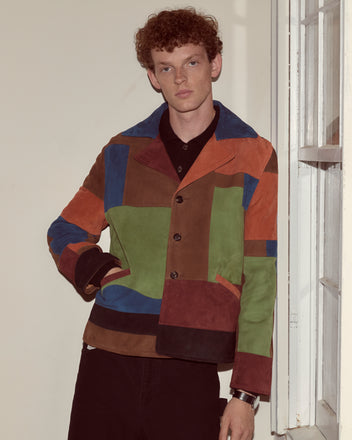 Person with curly hair wearing a Suede Collage Jacket stands by a window, looking at the camera.