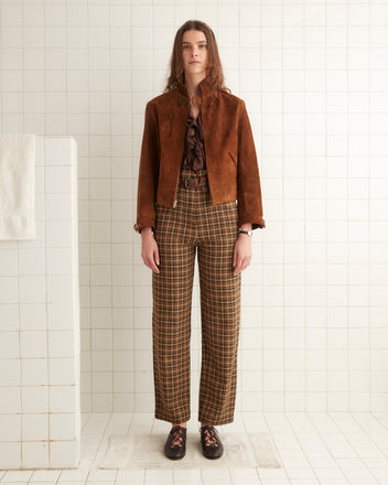 A person with long hair stands in a tiled room, wearing a dark brown Suede Howard Jacket, a frilled blouse, plaid trousers, and black shoes. A white towel hangs on a tiled partition to the left.