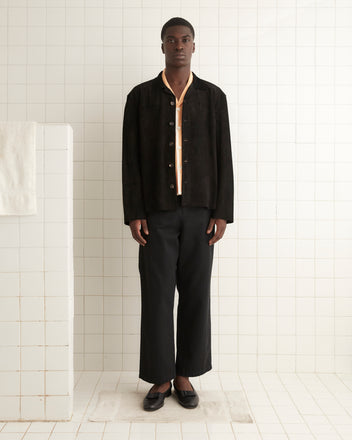 A man stands in a tiled room wearing a Suede Overshirt - Black over a yellow shirt, reminiscent of a 1960s-1970s work shirt, dark trousers, and black shoes. He stands on a white rug next to a white half wall with a towel draped over it.