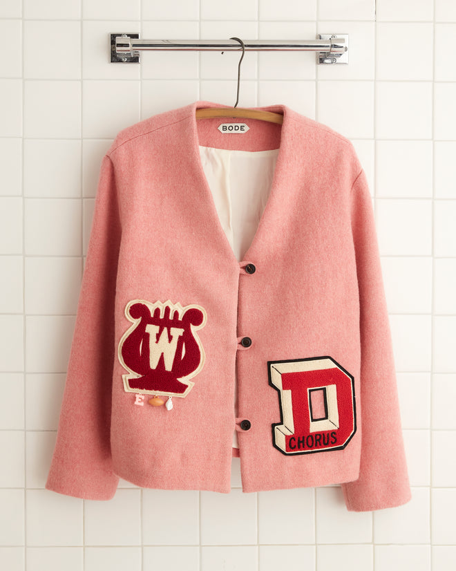 The Sugarplum Jacket - 6, featuring a boxy fit and adorned with emblem patches, hangs on a white tile wall, exuding vintage charm in its pink varsity style.