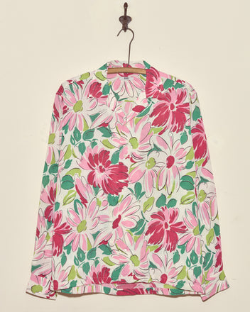 The Sukhothai Flower Shirt is a long-sleeve vintage top with a bold pink, white, and green floral print, displayed on a wall hook.