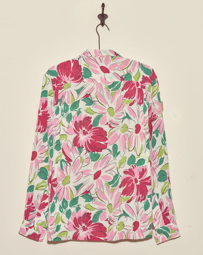 The Sukhothai Flower Shirt, a long-sleeve vintage top with a vibrant pink, white, and green floral pattern, hangs on a hook against a cream wall.