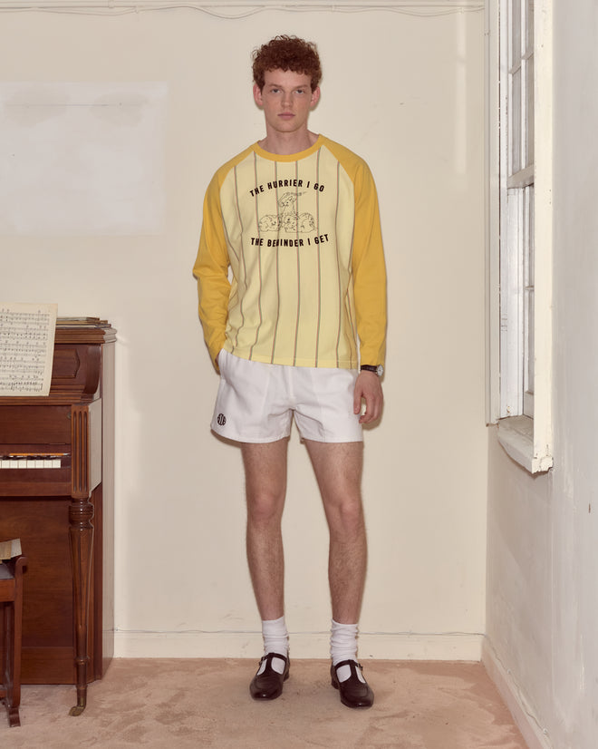 A man in a Summertime Stripe Baseball Tee and white shorts stands indoors by a piano and a large window.