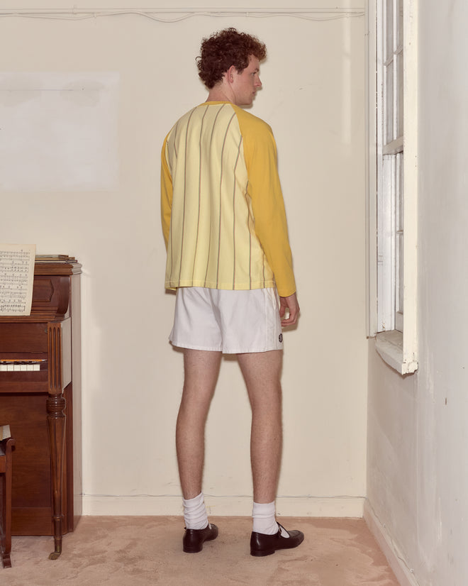 A man in a Summertime Stripe Baseball Tee and white shorts stands indoors, facing away from the camera near a piano and window.