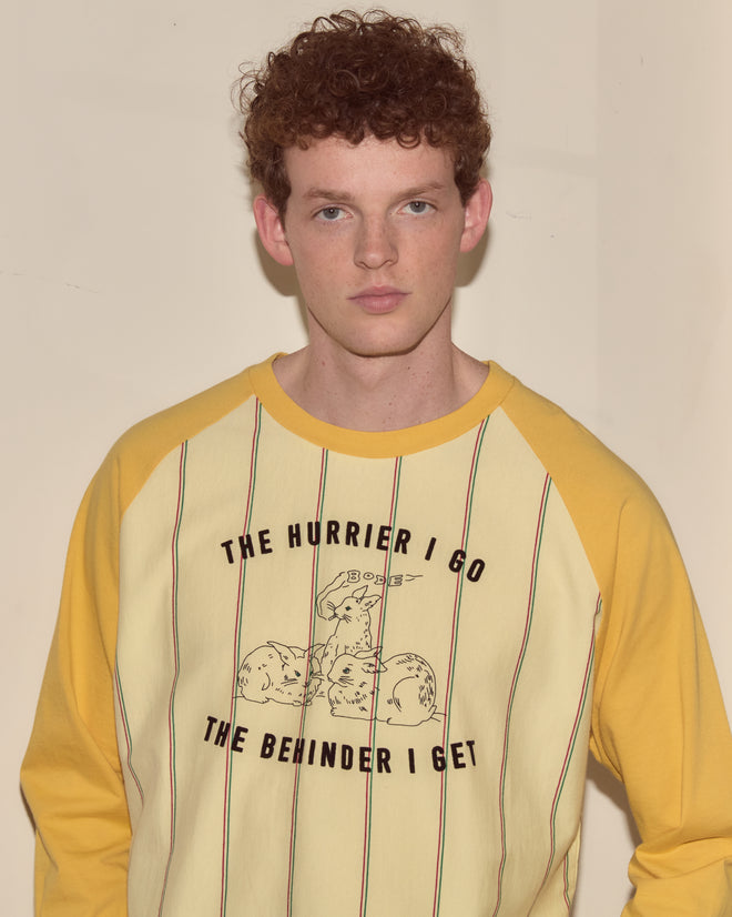 A young man with curly red hair wears the Summertime Stripe Baseball Tee in yellow and cream with the text "THE HURRIER I GO THE BEHINDER I GET.