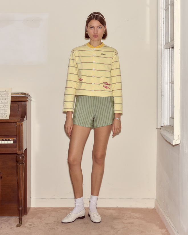 A woman stands indoors by a piano and window, wearing the Summertime Stripe Cardigan in yellow stripes with green shorts.