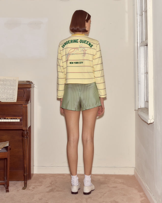 A woman stands indoors near a piano and window, facing away and wearing the Summertime Stripe Cardigan over a striped top with shorts.