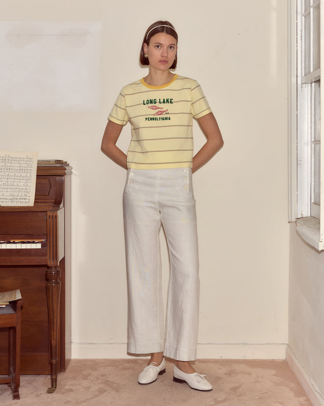A woman in the Summertime Stripe Tee and white pants stands indoors by a piano and window, conjuring memories of Camp Onibar by Long Lake.
