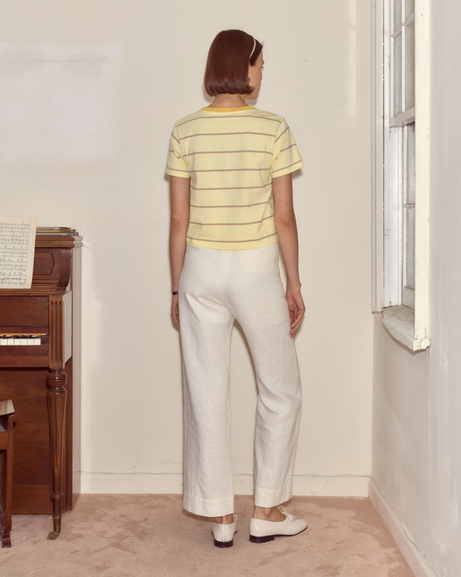 A person with short hair, wearing the Summertime Stripe Tee and white pants, stands facing away in a room with a piano.