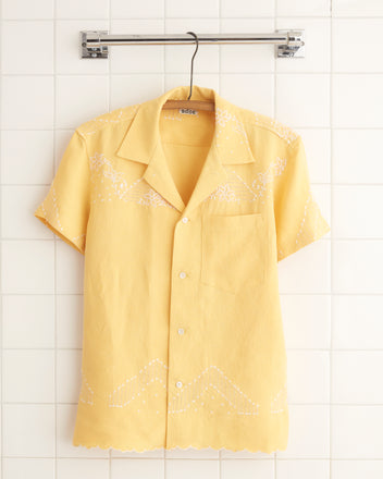 The Sun Flare Shirt - S, a yellow short-sleeved embroidery shirt with white stitching, hangs on a hanger against a white tiled wall, evoking the charm of a midcentury tablecloth.