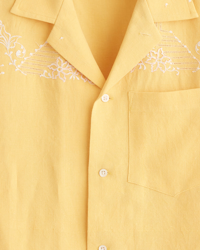 The Sun Flare Shirt - S, a yellow short-sleeved embroidery shirt with white stitching, hangs on a hanger against a white tiled wall, evoking the charm of a midcentury tablecloth.