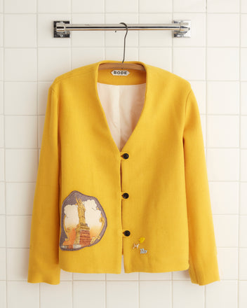 The Sunsoaked Blanket Jacket - Size 2 exudes a vintage charm with its yellow hue, patch detailing, and three-button design, all displayed stylishly against a tiled wall. Its boxy fit enhances its retro allure, harking back to the nostalgia of a midcentury wool blanket.