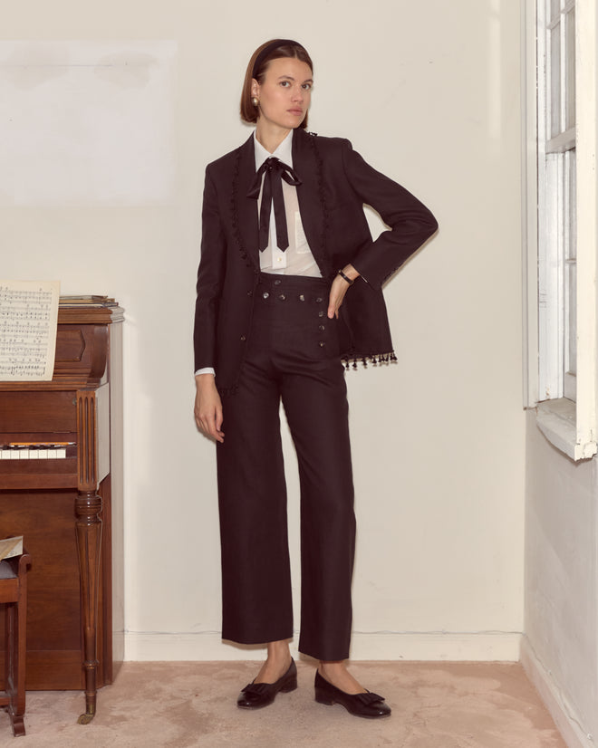 A woman in the Surrey Fringe Jacket, featuring a black linen shawl collar, pairs it with a white shirt and bow tie as she stands by a piano in a softly lit room.