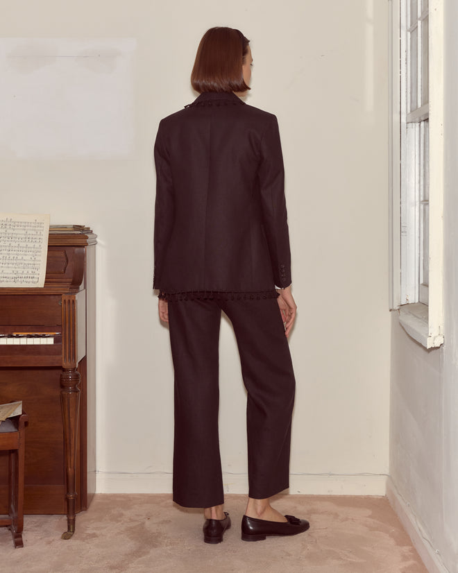 Wearing the Surrey Fringe Jacket, a person stands with their back turned by a piano and window in a minimalist room. Subtle beaded fringe adds elegance to the black linen shawl-collar silhouette.