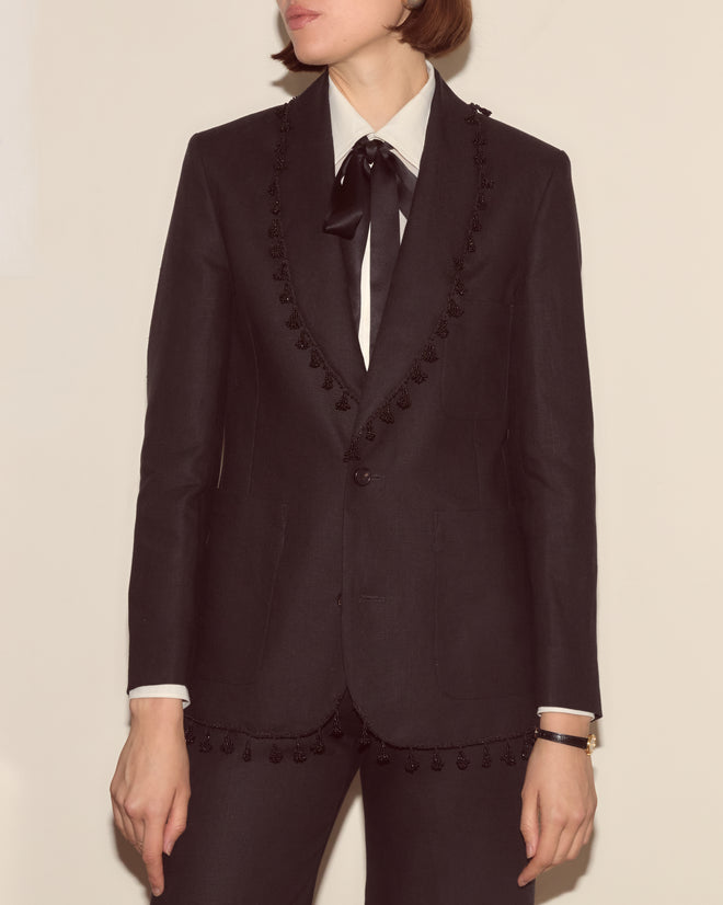 Person wearing the Surrey Fringe Jacket—a black linen shawl-collar jacket with decorative beaded fringe—paired with a white shirt, black tie, and wristwatch, standing against a plain wall.