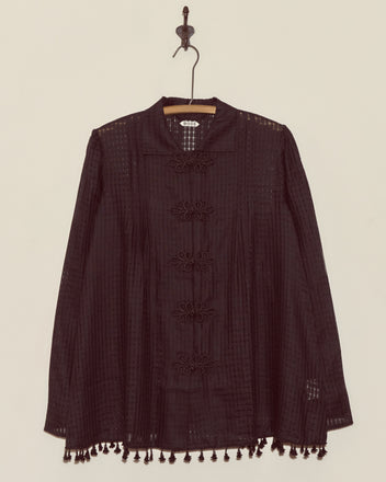 The Surrey Fringe Shirt, a black long-sleeve blouse with gauzy fabric and tassels on the hem, hangs on a wooden hanger against a white wall.