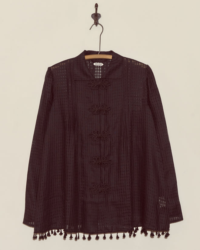 The Surrey Fringe Shirt, a black long-sleeve blouse with gauzy fabric and tassels on the hem, hangs on a wooden hanger against a white wall.