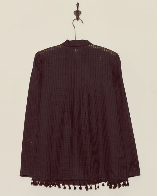 The Surrey Fringe Shirt is a gauzy black long-sleeve blouse with tassels at the hem, displayed on a wall hook.
