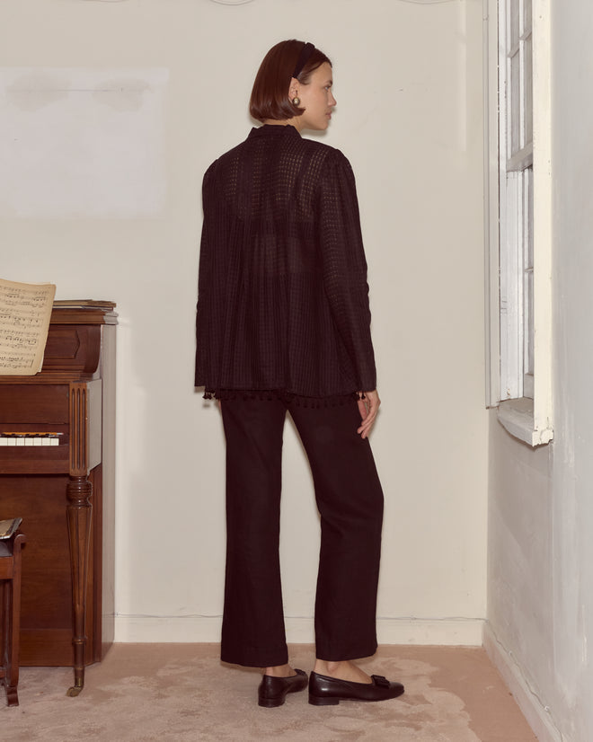 A woman in black clothes with a gauzy Surrey Fringe Shirt stands indoors, facing away, beside a piano and a window.