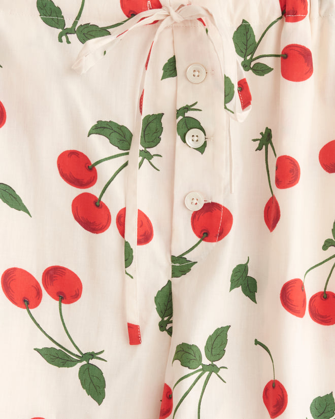 The Sweetcherry Pajama Pants - XS, featuring a delightful cherry print, hang on a wooden hanger against a white tiled wall. Made from deadstock yardage, these cozy pants infuse vintage charm into your bedtime routine.