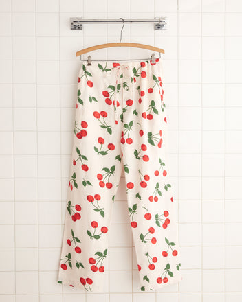 The Sweetcherry Pajama Pants - XS, featuring a delightful cherry print, hang on a wooden hanger against a white tiled wall. Made from deadstock yardage, these cozy pants infuse vintage charm into your bedtime routine.