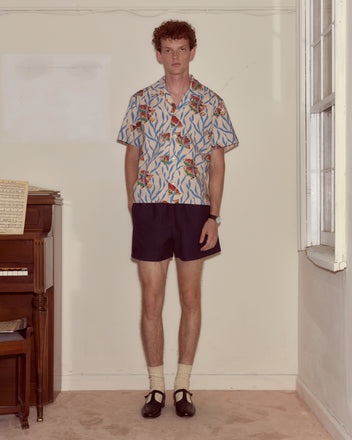 A man wearing the Swimmers Short Sleeve Shirt with a blue seaweed pattern stands indoors by a piano, looking at the camera.