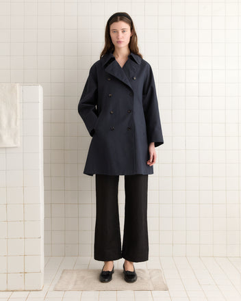 A woman in a dark Waxed Herringbone Peacoat and pants stands with hands in pockets on a white-tiled floor, against a tiled wall.