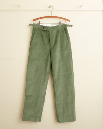 Sycamore Corduroy Trousers with side buckle waist adjusters hanging on a wooden hanger against a light-colored wall.