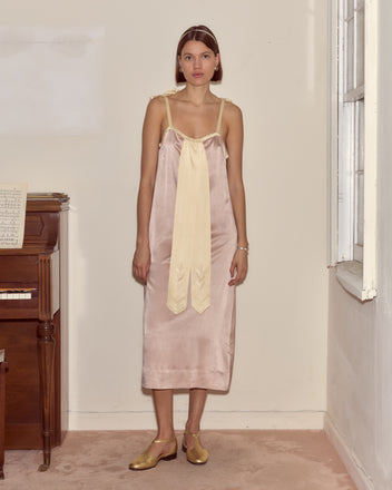A woman stands indoors by a piano and window, wearing the Sylphid Dress—a lilac pink silk gown adorned with a large cream bow.