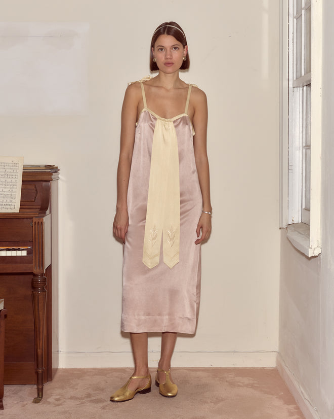 A woman stands indoors by a piano and window, wearing the Sylphid Dress—a lilac pink silk gown adorned with a large cream bow.