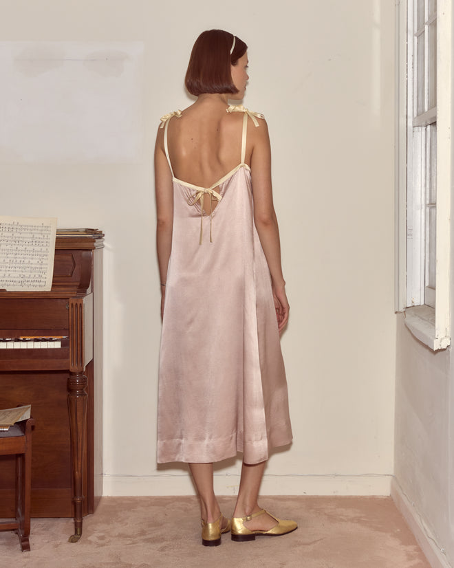 A woman in the Sylphid Dress, a lilac pink silk gown, stands by a piano in a softly lit room, facing away.