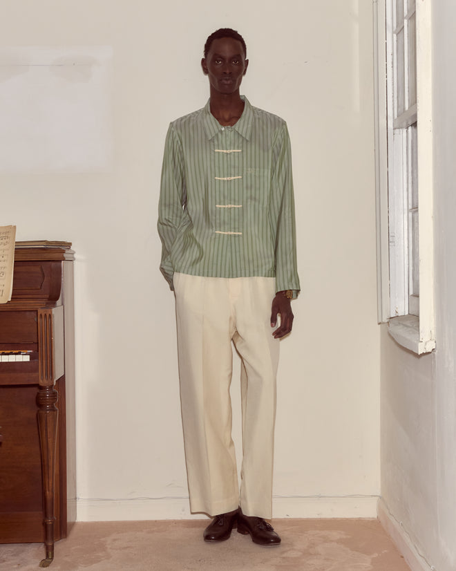 Indoors by a piano and window, a person wears cream trousers and the Sylvan Stripe Long Sleeve Shirt.