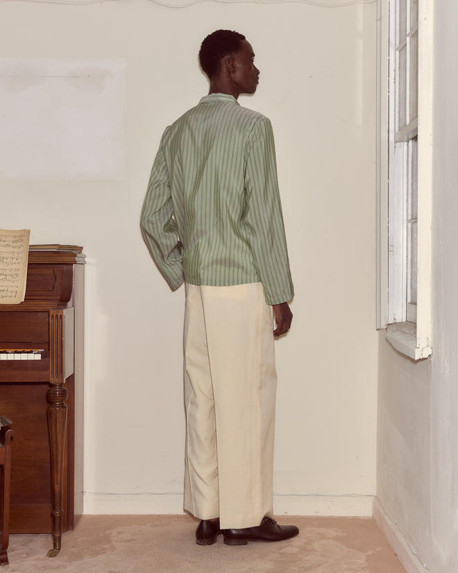 A person in a Sylvan Stripe Long Sleeve Shirt and cream pants stands facing a wall near a window and an upright piano.