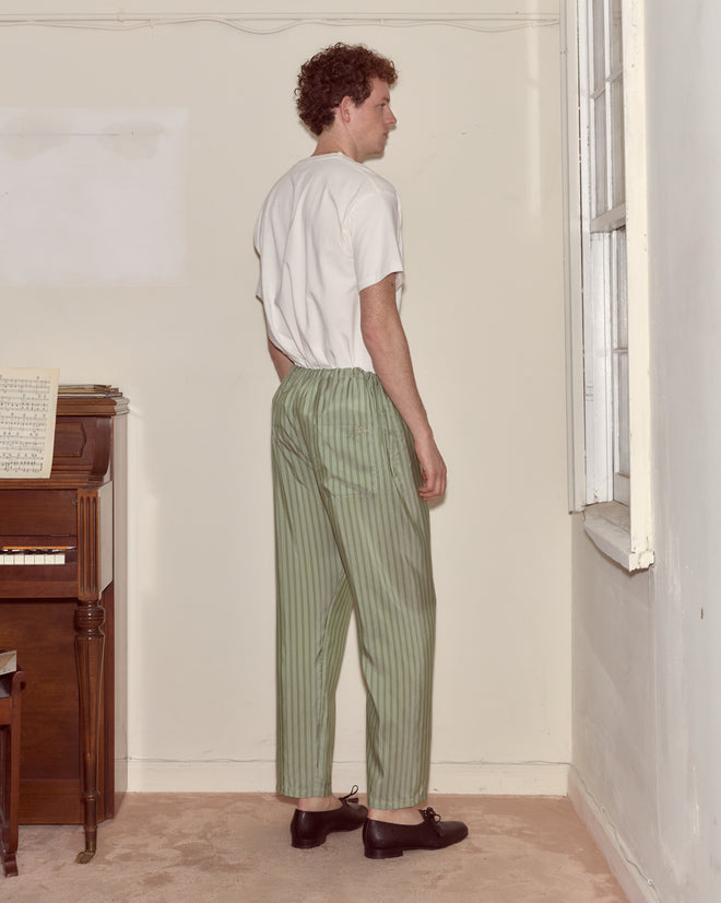 A man with curly hair and a white shirt wears Sylvan Stripe Pajama Pants in a classic green striped design, standing by a window with his back to the camera.
