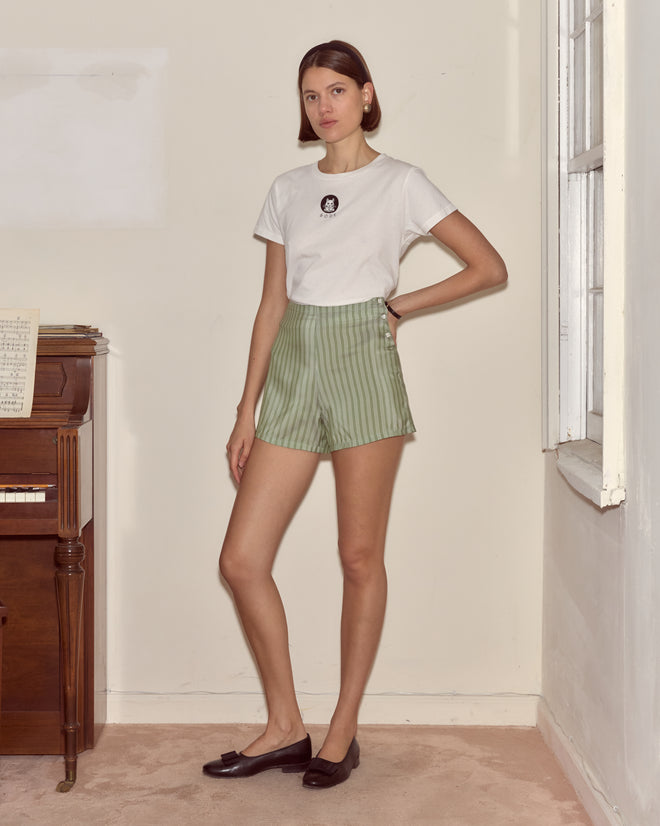 Woman in a white t-shirt and Sylvan Stripe Shorts - Green stands indoors near a piano and a window.