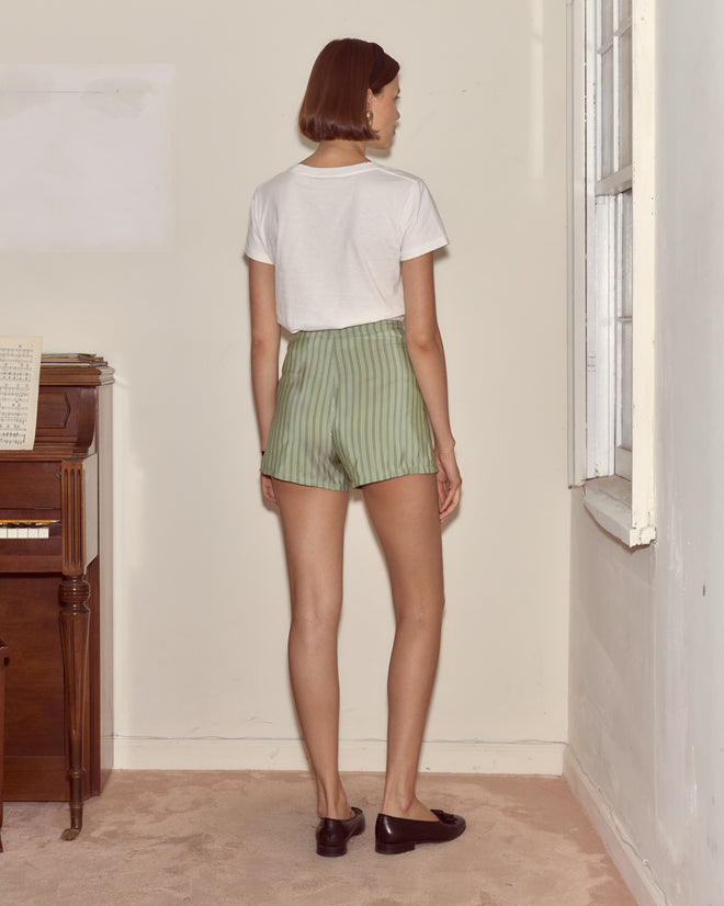 A woman stands indoors near a piano and window, facing a wall, wearing a white shirt and Sylvan Stripe Shorts - Green.