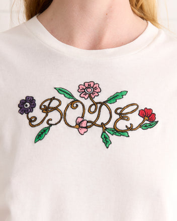 The Posy Tee is a white shirt made from 100% cotton, showcasing a vibrant floral pattern that spells 