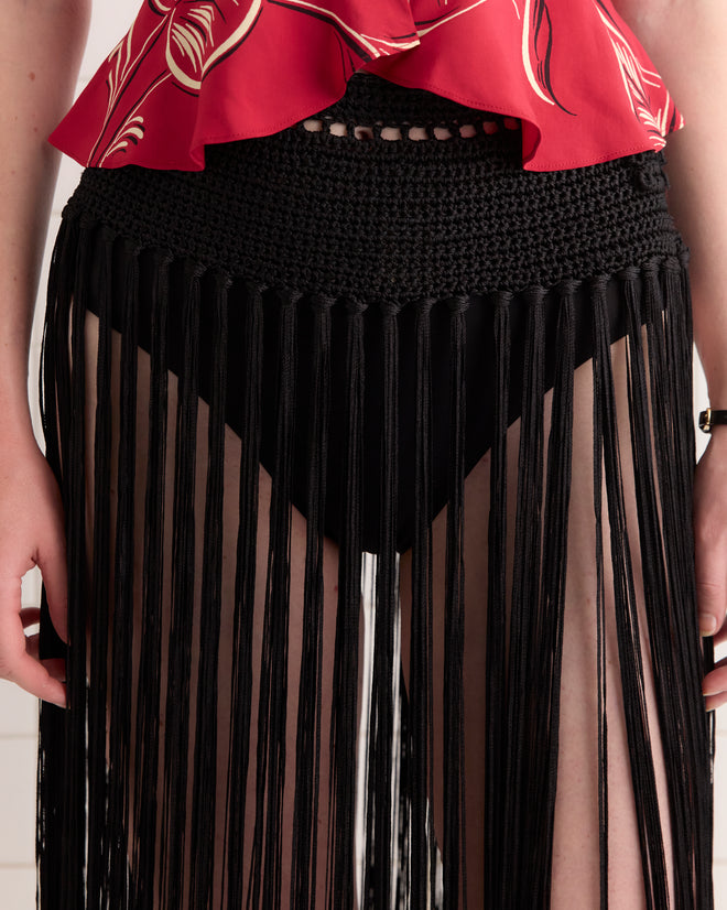 Person wearing a red top and the Crochet Fringe Skirt - Black, a vintage-style skirt with long fringe strands over black bottoms.