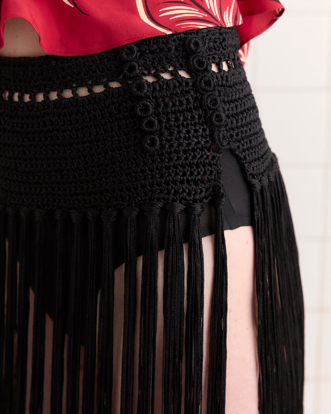 Person wearing a red top and the Crochet Fringe Skirt - Black, featuring a vintage crochet design with long fringe details over black shorts.