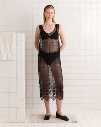 A woman wears the Beaded Grid Vine Dress - Black, a sheer black silk crepe dress with a geometric vine pattern, and black flats while standing in a white tiled room with a towel hanging on the wall.