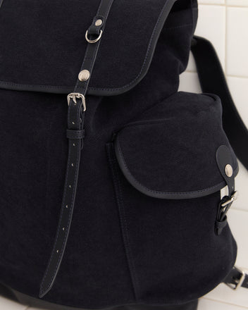 Close-up of the Canvas Rucksack - Black, crafted from heavyweight canvas with leather accents and a small side pocket, set against a tiled wall.