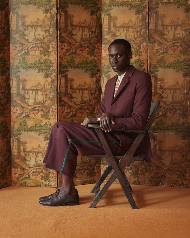 A person in a maroon suit, paired with Riding Twill Braided Trousers, sits on a wooden chair against a vintage wallpaper backdrop.