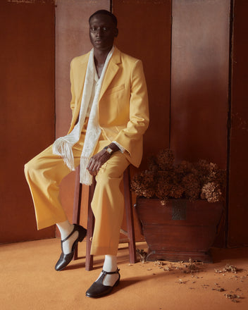 A person in a Riding Twill Suit Jacket - Yellow, paired with a white scarf, sits on a stool against a brown background adorned with dried flowers, the ensemble's pearl buttons subtly catching the light.