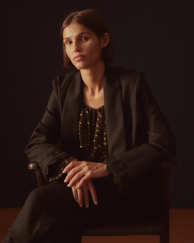 A person stands against a light-colored wall wearing the Ripple Jacket, a black suit featuring scalloped edges and rickrack trim, elegantly paired with black shoes.