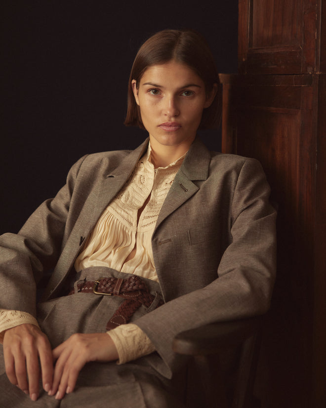 A person wearing the Lillian Suit Jacket, crafted from a tailored brown wool/linen blend and paired with a white shirt, stands in front of a cream-colored wall on a patterned carpet.